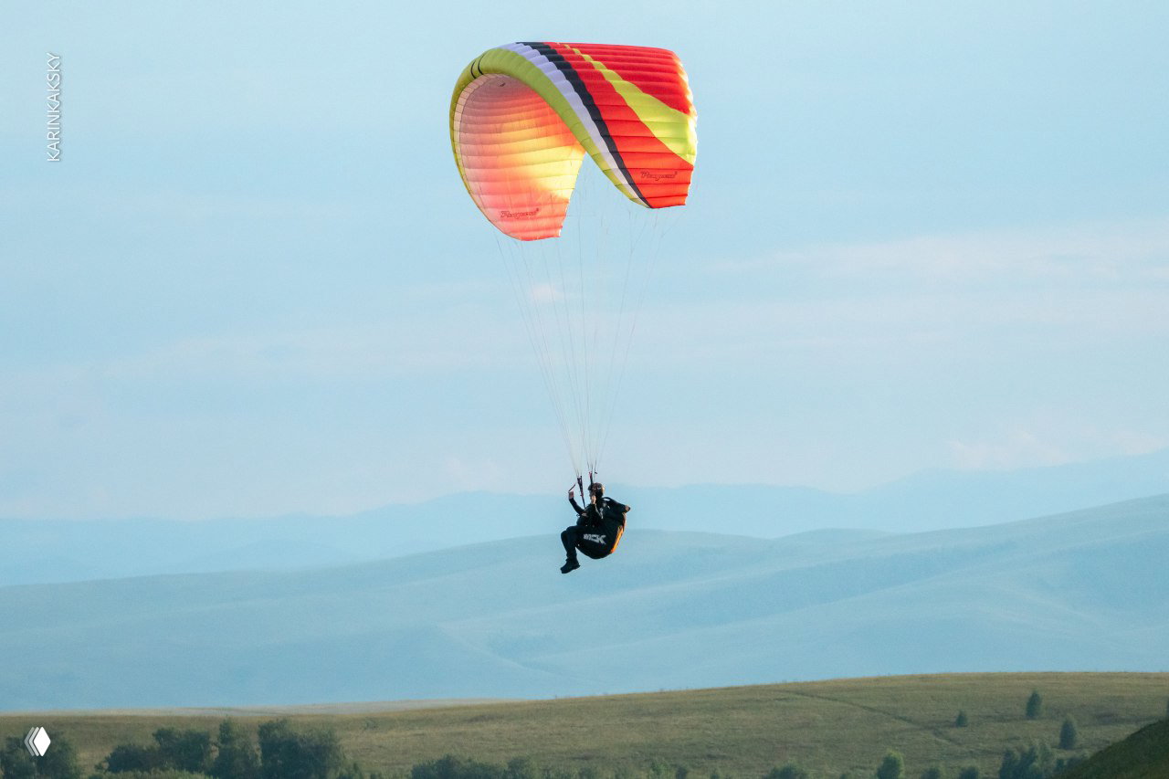 Пилот на красно‑жёлтом крыле парит над полями и холмами, видна дистанция до берега и плавные линии ландшафта.