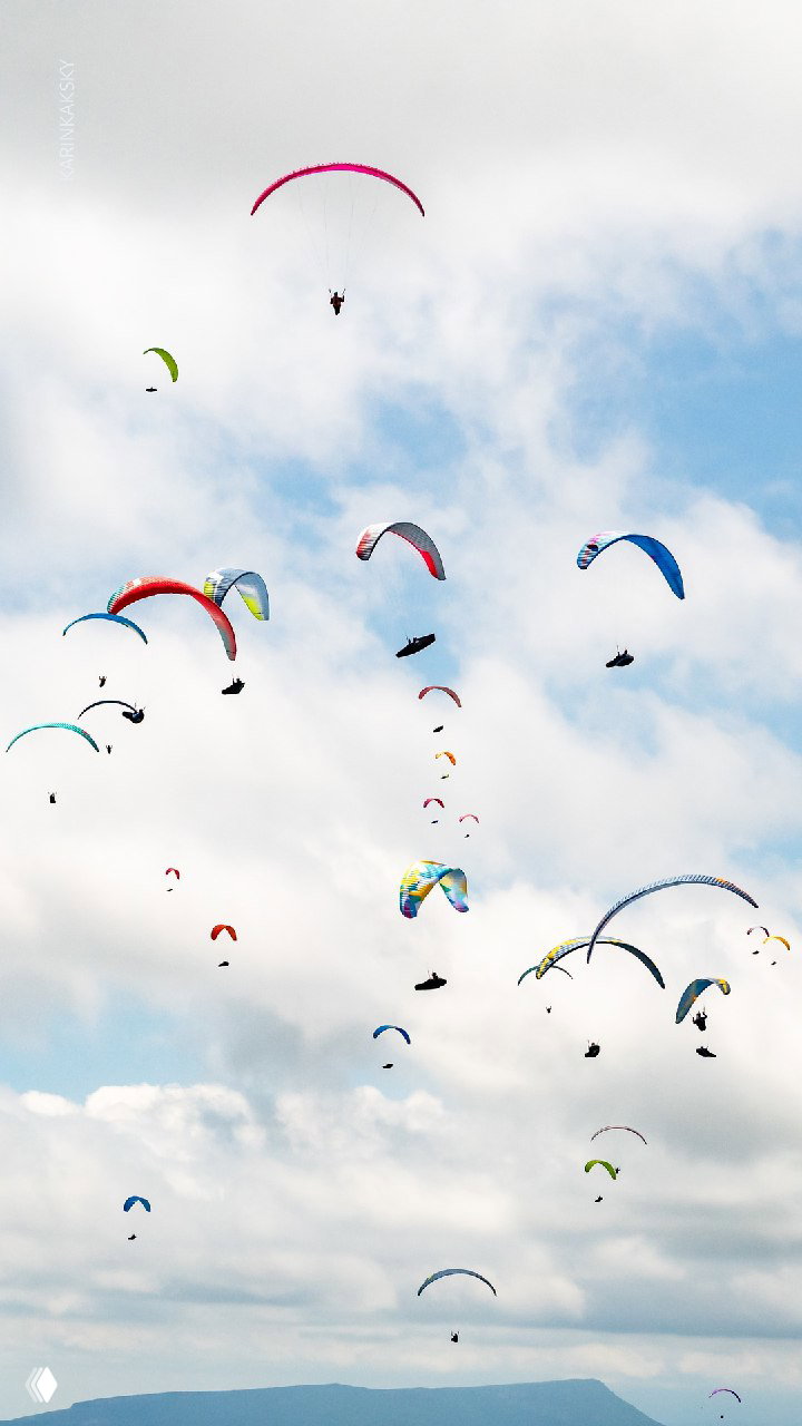 Многоцветные парапланы под облачным небом: множество пилотов в воздухе на фоне рассеянных облаков и большого простора для полёта.