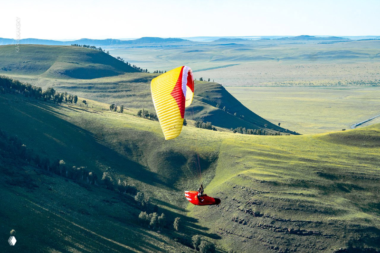 Жёлто-красный параплан над просторной степью и холмами Хакасии: один пилот на фоне широкого пейзажа и мягких склонов.