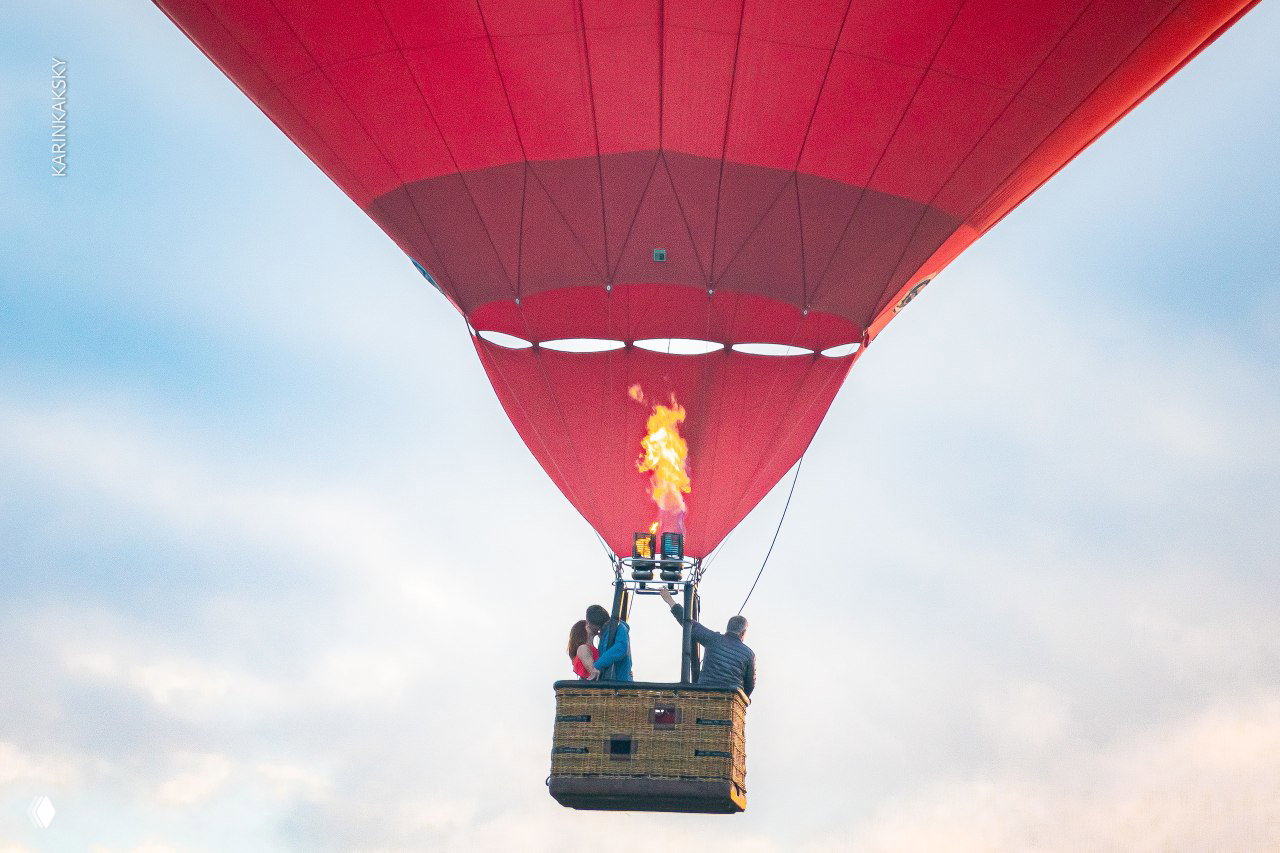 Вид сзади на красный аэростат в полёте: корзина с людьми, горелка и форма купола шара на фоне светлого неба и облаков.