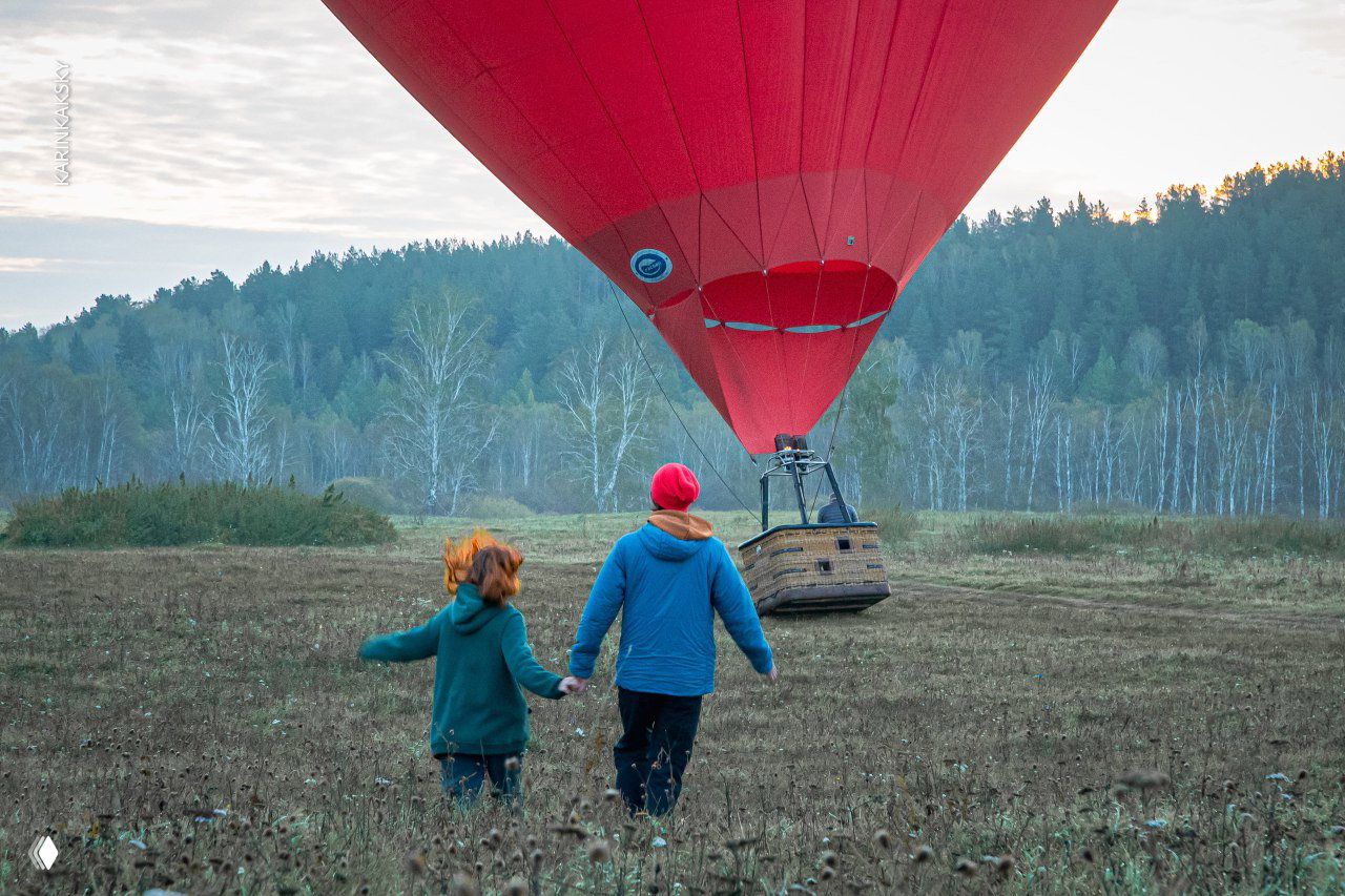 Красный воздушный шар садится на поле: два человека держатся за руки и идут к корзине на фоне лесного полотна и туманного утра.