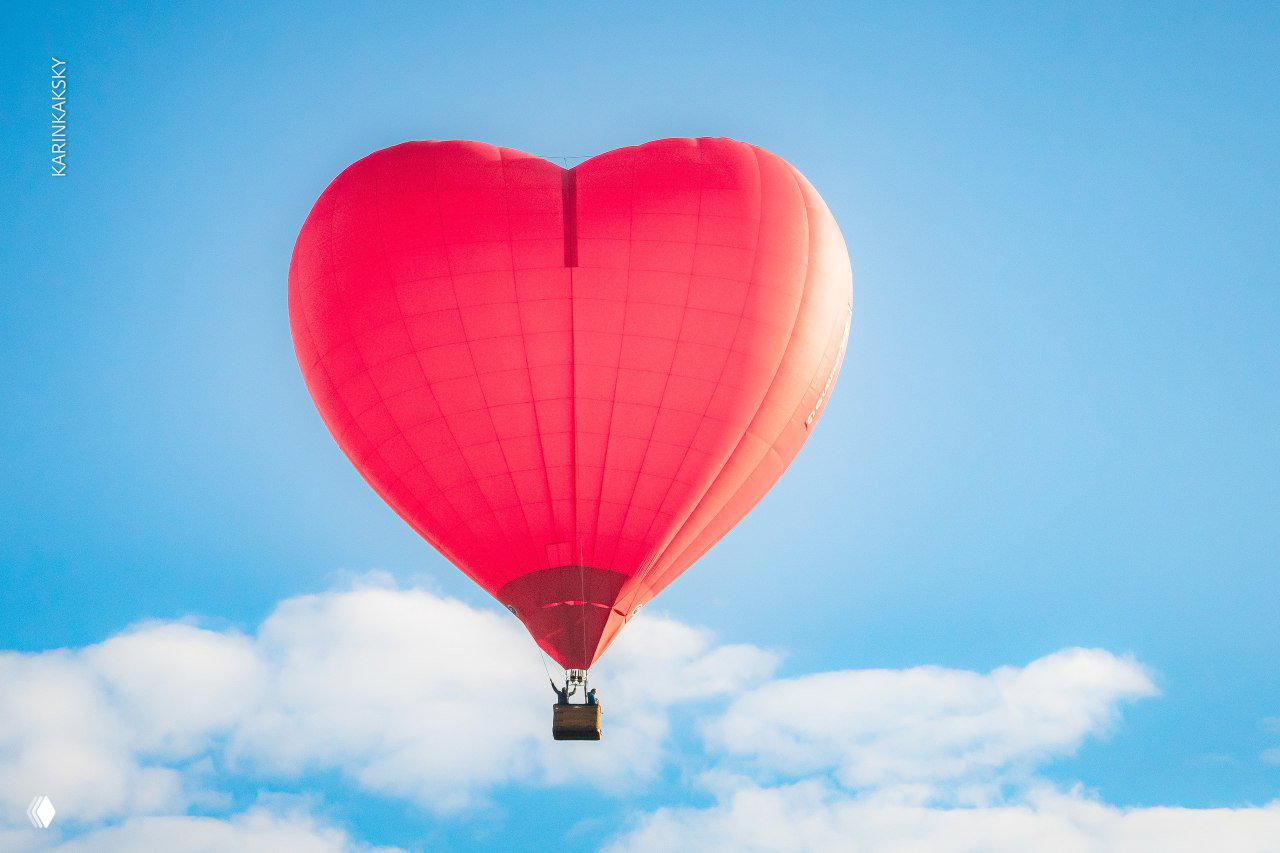 Форма шара в виде большого сердца ярко-красного цвета, парящий в голубом небе с лёгкими облаками и маленькой корзиной внизу.