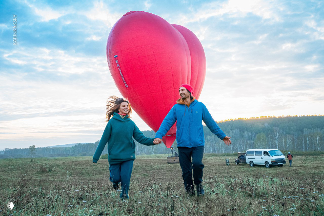 Пара идёт по полю, держась за руки, на фоне надутого красного шара; образ радости и лёгкости после полёта в естественном пейзаже.