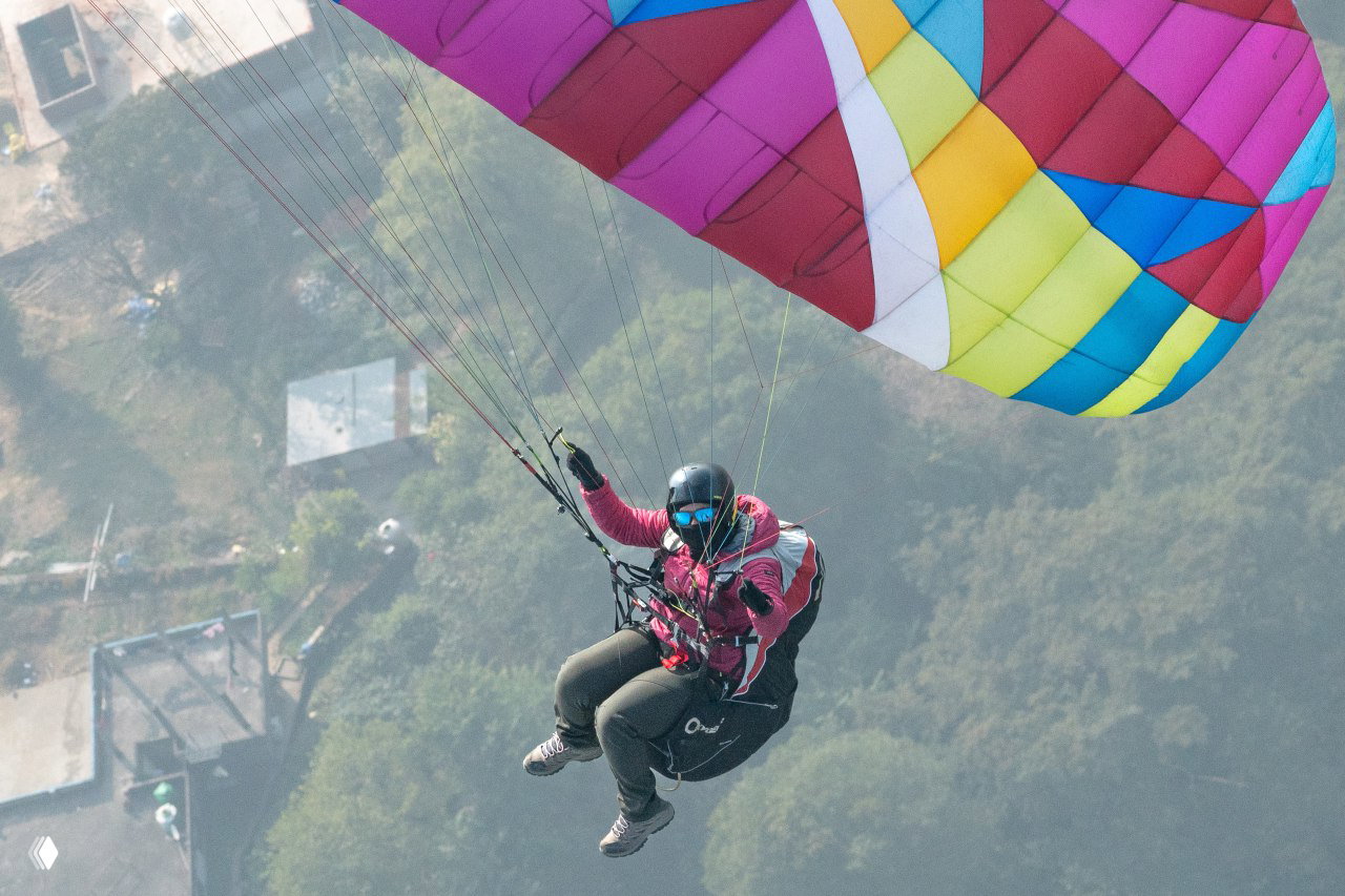 Крупный план пилота в тёплой экипировке под ярким многоцветным крылом параплана, полёт с выразительной перспективой и акцентом на эмоции в воздухе.