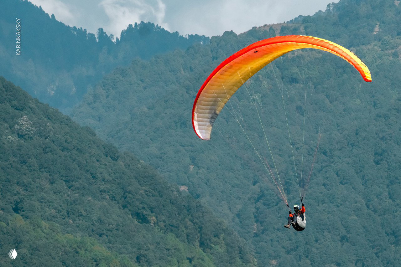 Тандемный параплан с двумя пилотами парит над зелёными предгорьями Индии; на склонах видны густые леса и ровные террасы.