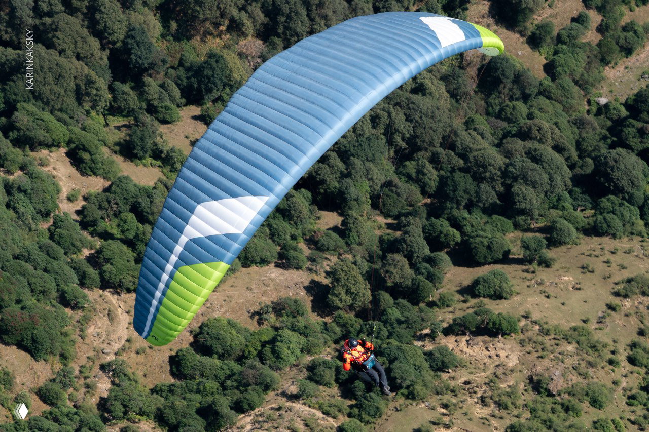 Голубое крыло параплана в полёте над кустарниковой горной местностью Индии; под крылом просматривается рельеф и тени от самолёта.