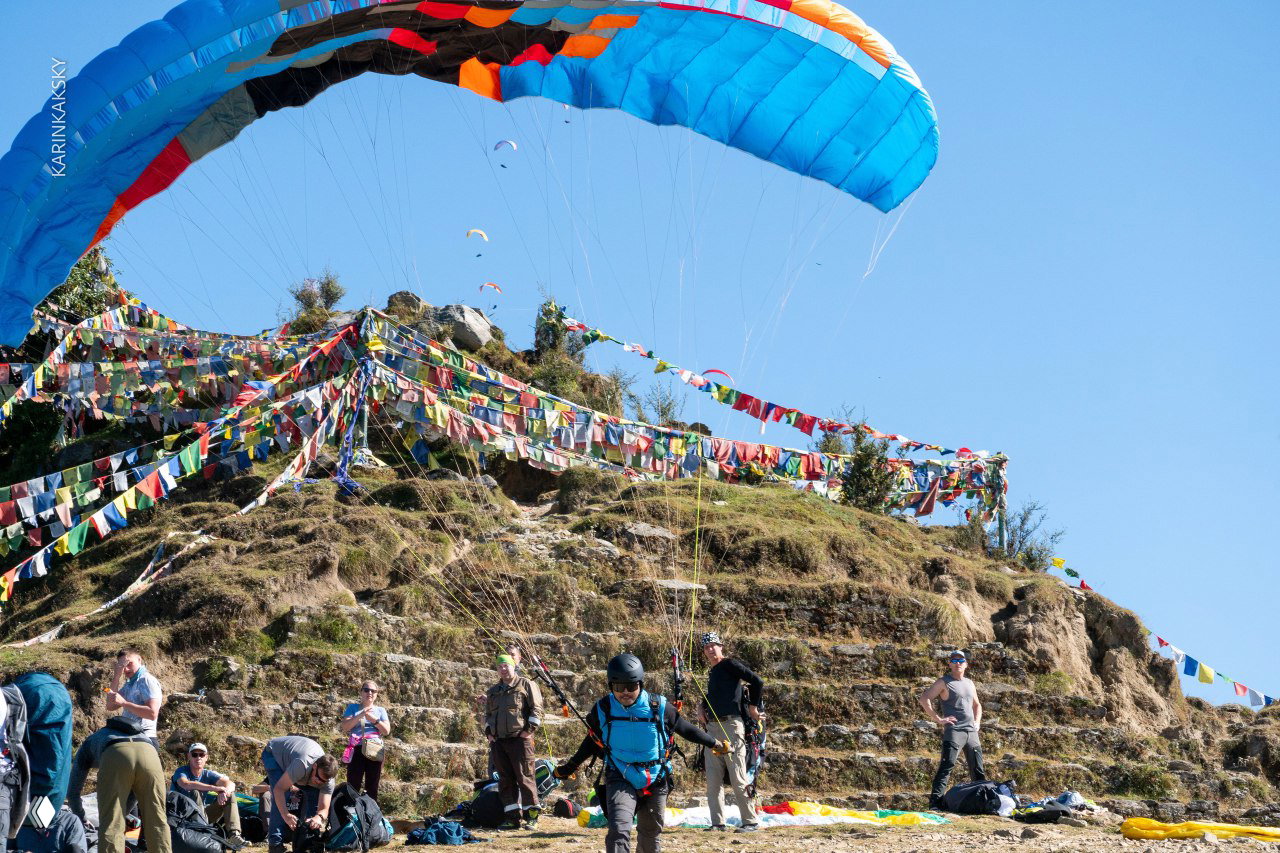 Стартовый холм с цветными молитвенными флажками, парапланом и группой людей — момент подготовки команды к полётам в Индии.