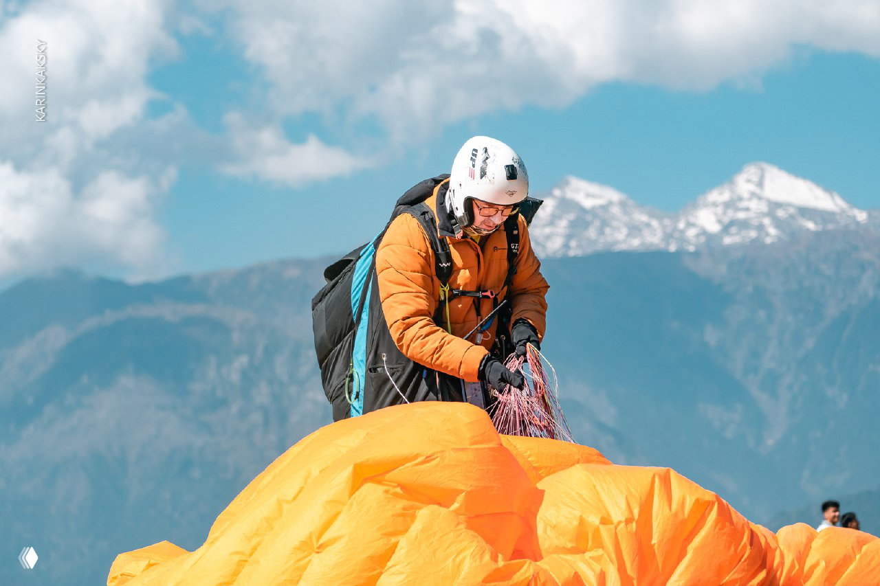 Пилот в оранжевой куртке проверяет стропы и купол параплана на фоне заснеженных гор — сцена подготовки к очередному взлёту.