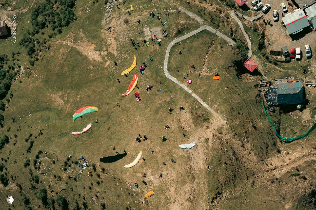 Аэрофотоснимок: разноцветные парапланы и группы людей на площадке старта в горах Индии; панорамный вид с высоты.