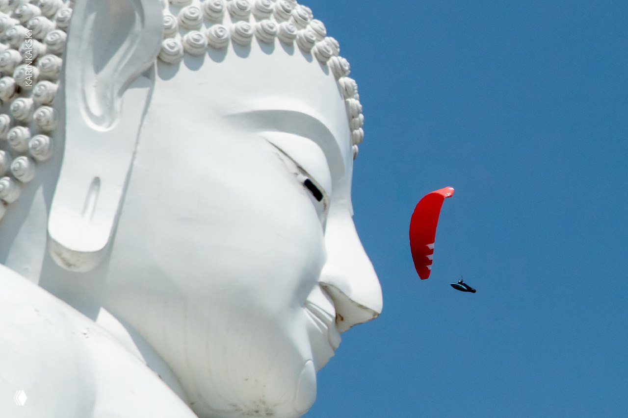 Красный параплан пролетает рядом с крупной белой статуей Будды — контраст религиозной архитектуры и воздушного спорта.