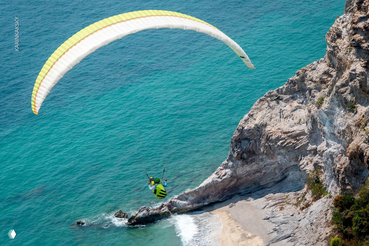 Параплан над бирюзовым морем и крутым побережьем: пилот в свободном полёте, пляж и прибрежный ландшафт на заднем плане.