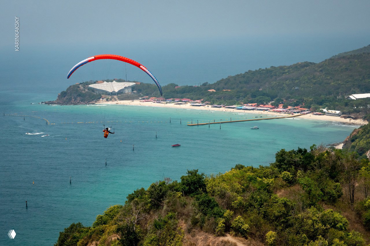 Параплан в небе над зелёным мысом и пляжем — широкая прибрежная панорама, море, берег и туристическая инфраструктура внизу.