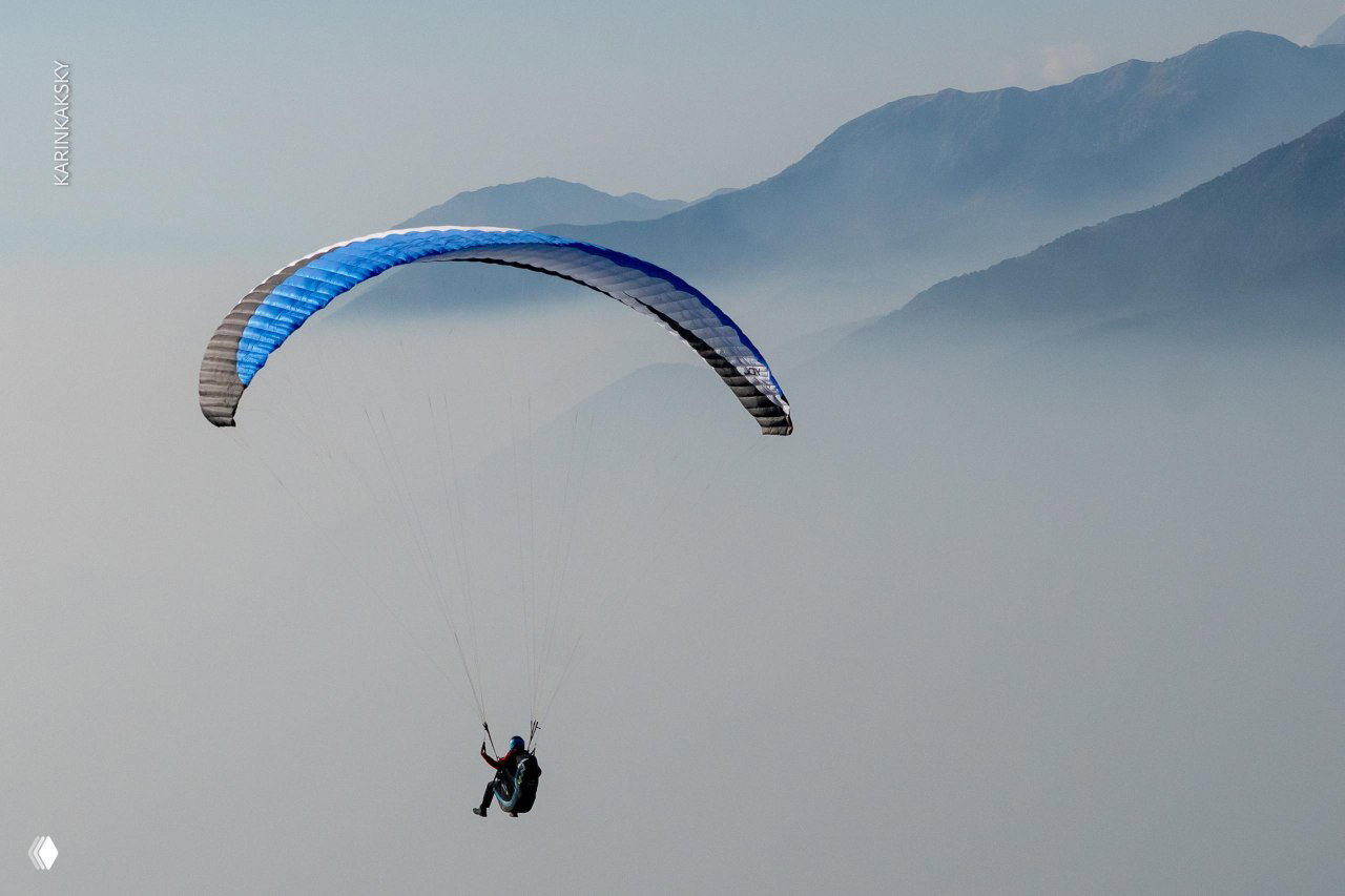 Одинокий пилот на синем крыле в мягком тумане: параплан парит на фоне горных силуэтов и лёгкой облачности Гималаев.