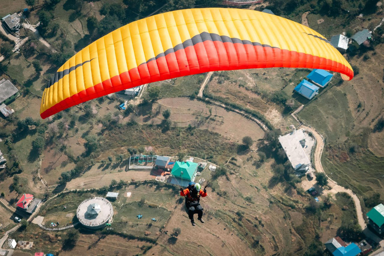 Пилот под жёлто-красным крылом летит над деревней и полями; кадр сверху показывает посадочную зону и местную застройку.