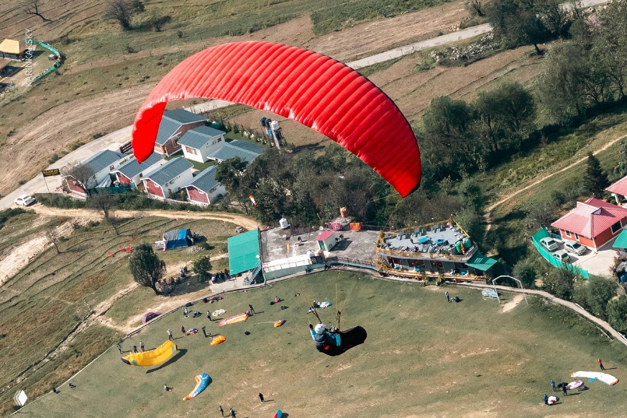 Красное крыло парит над зелёной площадкой; на земле видны раскладка парапланов и люди — сцена тренировочной сессии.