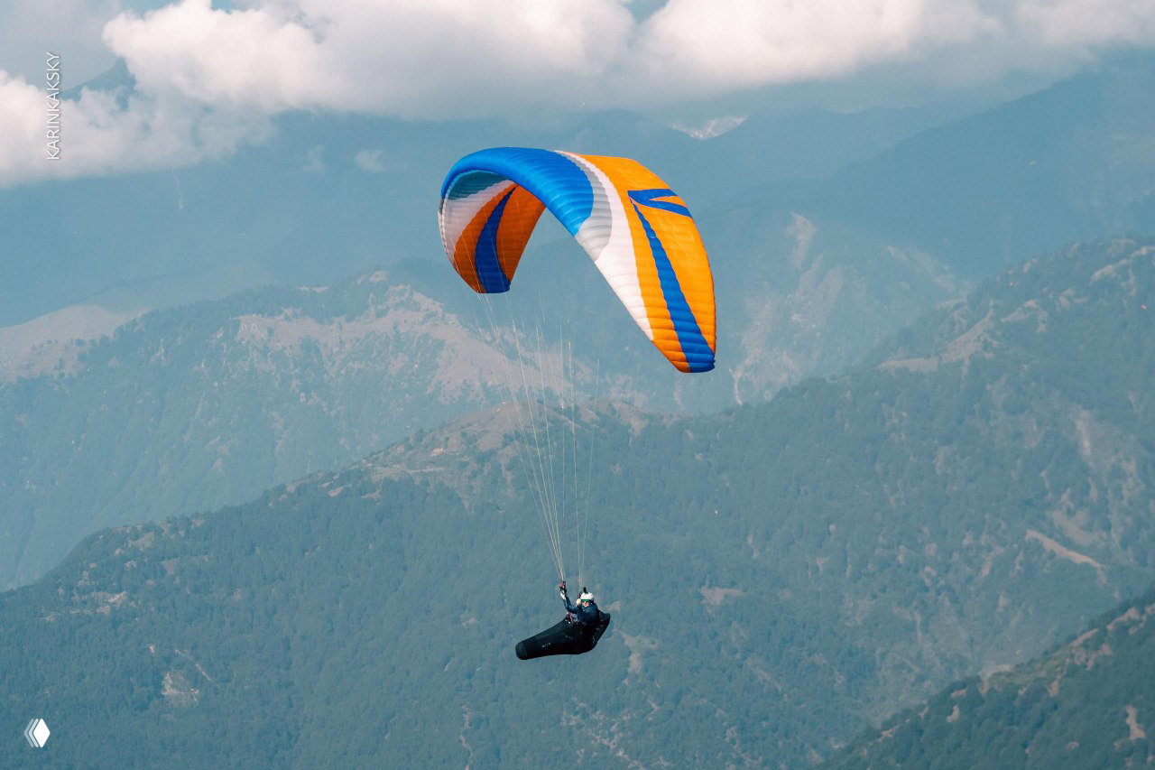 Яркое оранжево-синее крыло на фоне облаков и гор; пилот в подвесе держит курс вдоль гребня — атмосферный кадр из маршрута.