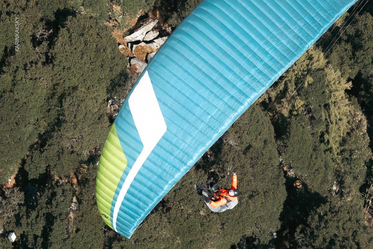 Крупный план голубо-зелёного крыла над скалистыми склонами; под ним виден пилот в комфортном подвесе — воздушная съёмка обучения.