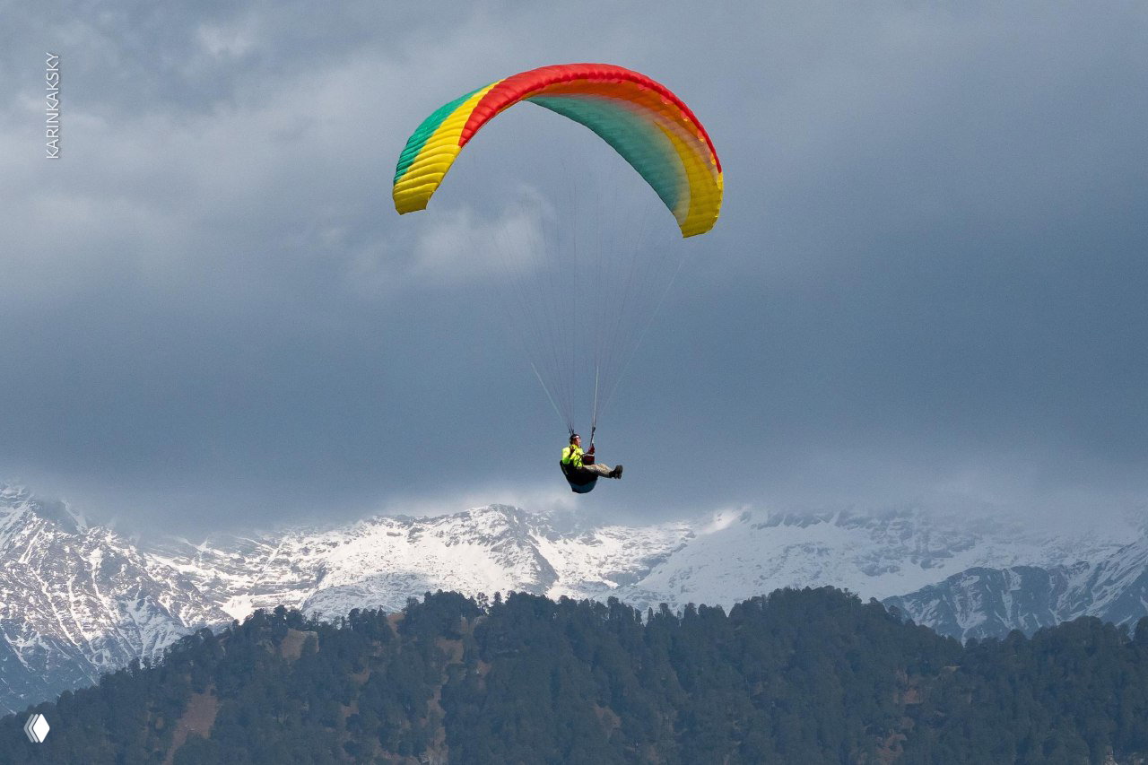 Радужное крыло в небе над лесистыми склонами и снежными вершинами на заднем плане; пилот выполняет полёт вдоль хребта Гималаев.