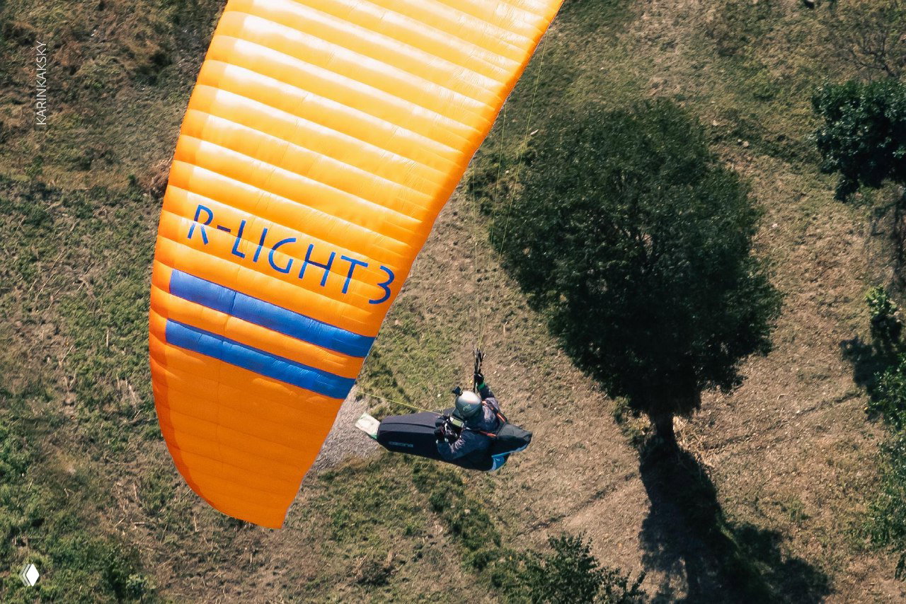 Оранжевое крыло и пилот видны сверху над травянистым ландшафтом и одиночным деревом — момент во время учебной сессии.