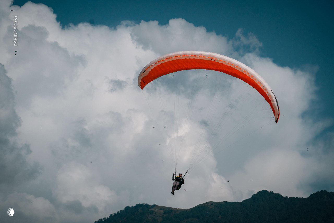 Парапланист под оранжево-белым крылом на фоне плотных облаков; кадр передаёт высоту и атмосферу учебного полёта в горах.