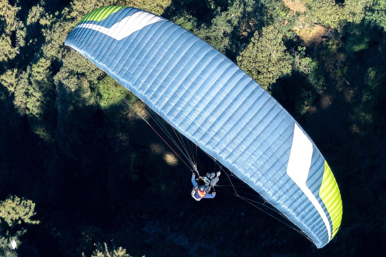 Крупный план синего параплана с пилотом, съёмка сверху показывает подвесную систему и контраст с тёмным лесным ландшафтом внизу.