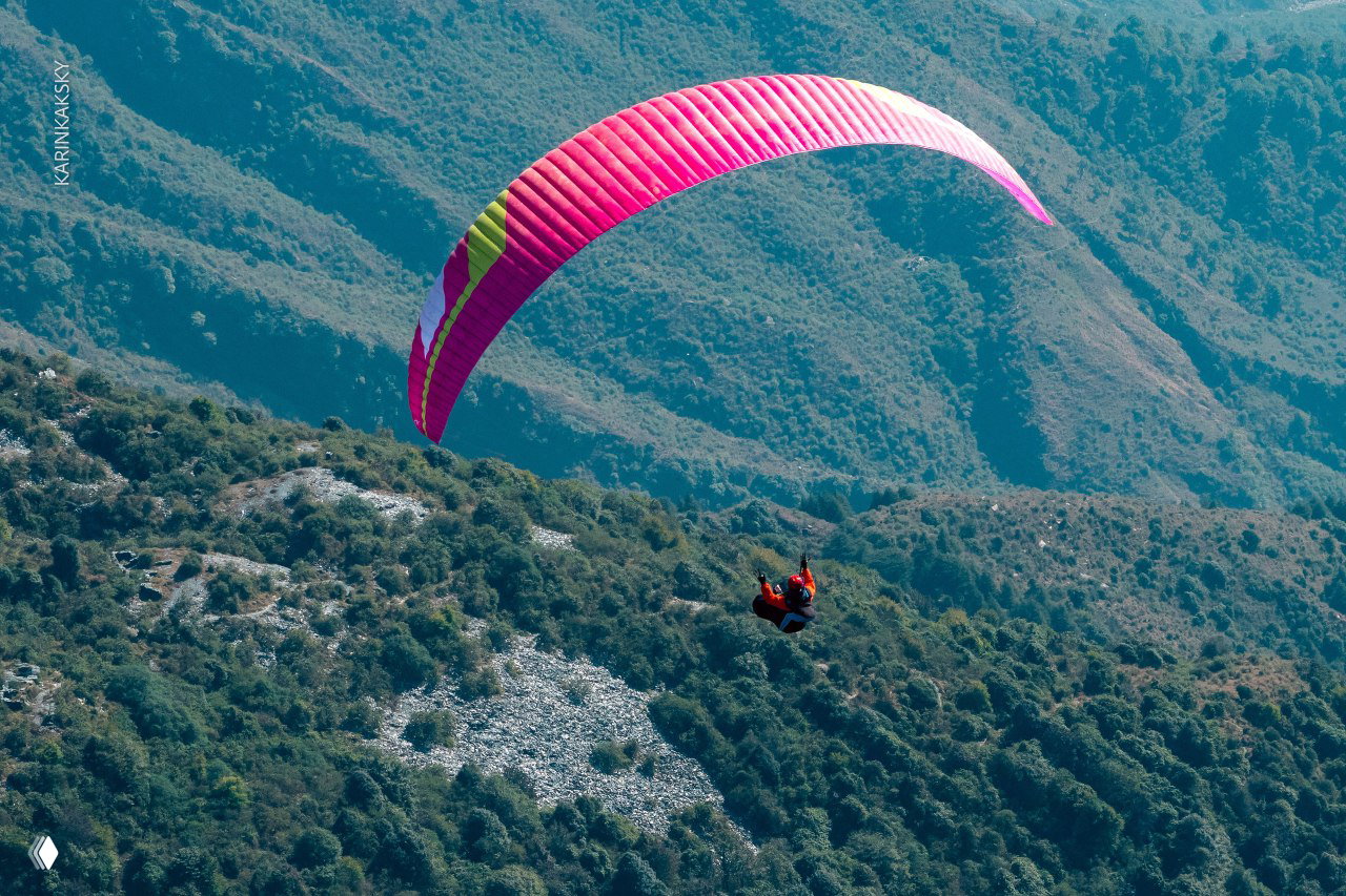 Розово‑фиолетовый параплан парит над зелёными холмами и скалистыми склонами, один пилот в подвеске на среднем расстоянии от камеры.