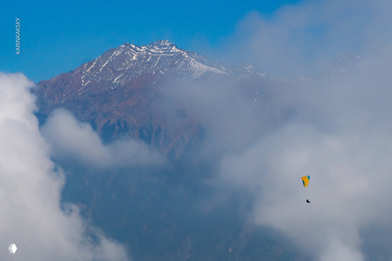 Небольшой параплан в облаках на фоне дальних заснеженных вершин; мягкие облачные слои подчёркивают глубину пейзажа.
