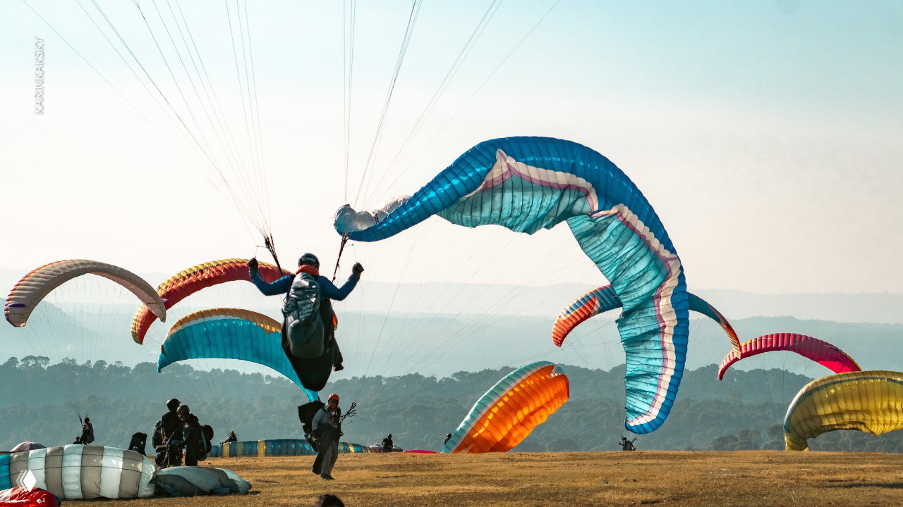 Группа парапланов и пилотов на стартовой поляне: развёрнутые крылья, подготовка экипажей и динамичная атмосфера перед вылетом.