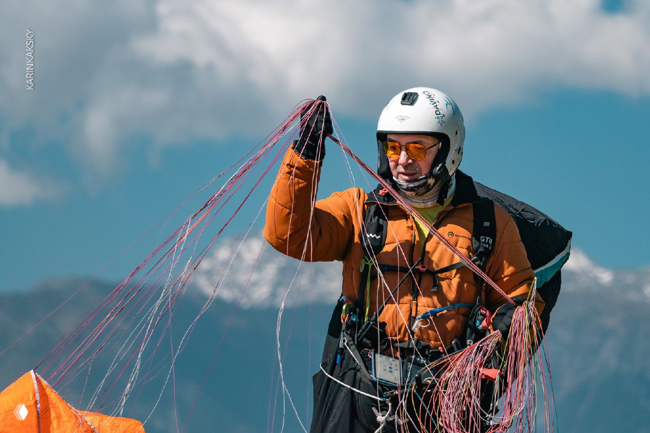Пилот в шлеме держит стропы параплана на фоне чистого неба и гор; детальный портрет подготовки перед полётом с видимыми линиями строп.