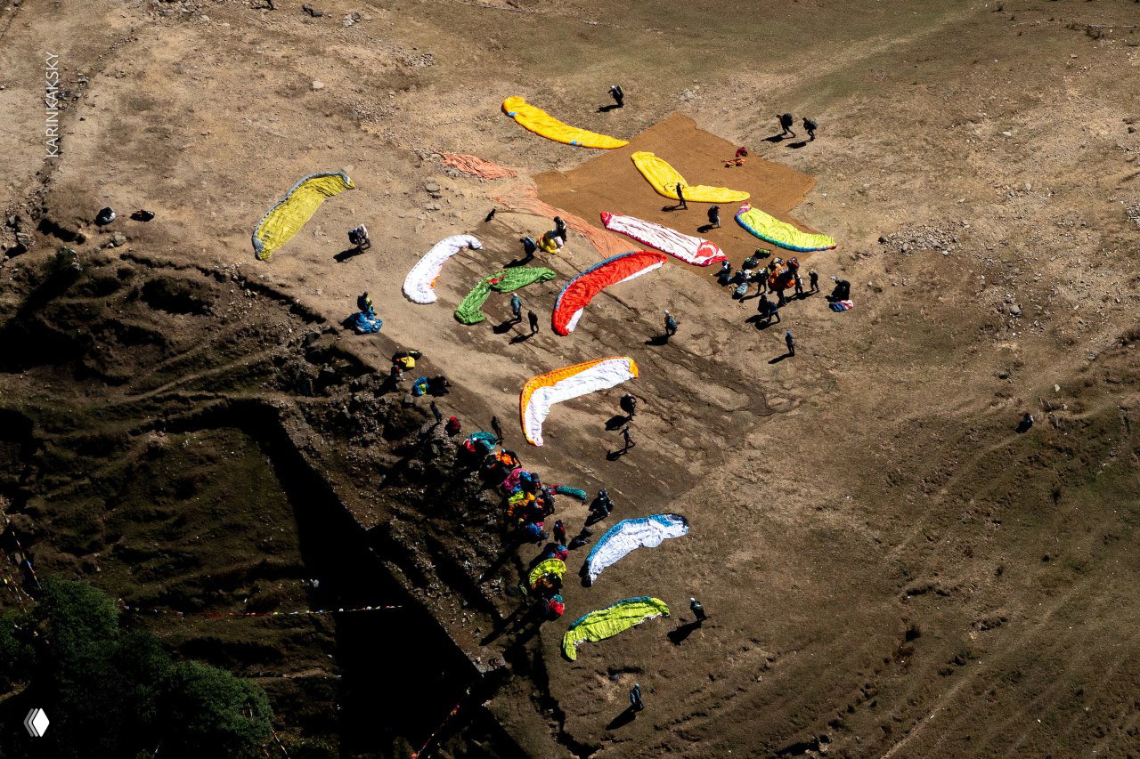 Аэросъёмка площадки со множеством разноцветных парапланов, разложенных на земле; видно организованную командную работу и масштаб события.