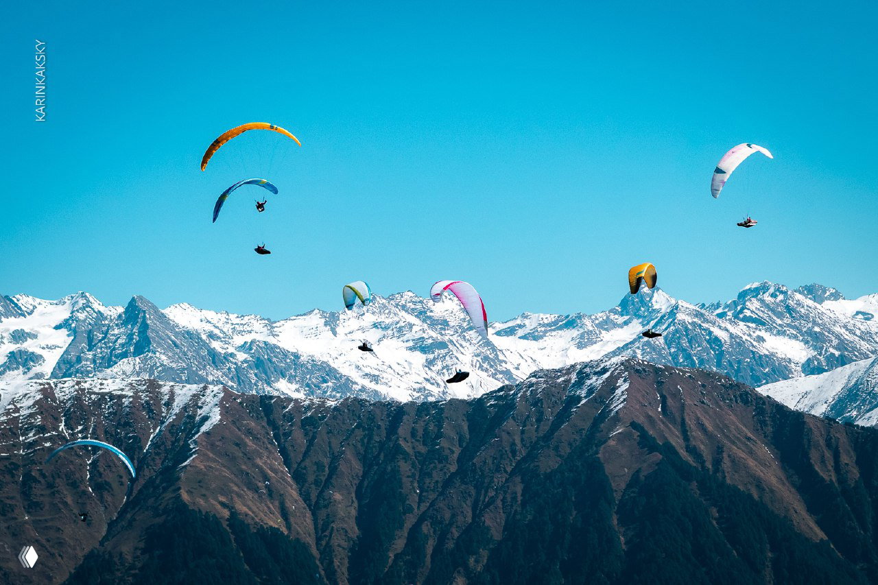 Несколько парапланов в небе над заснеженными хребтами; панорамный кадр подчёркивает масштаб полёта и простор горного пейзажа.