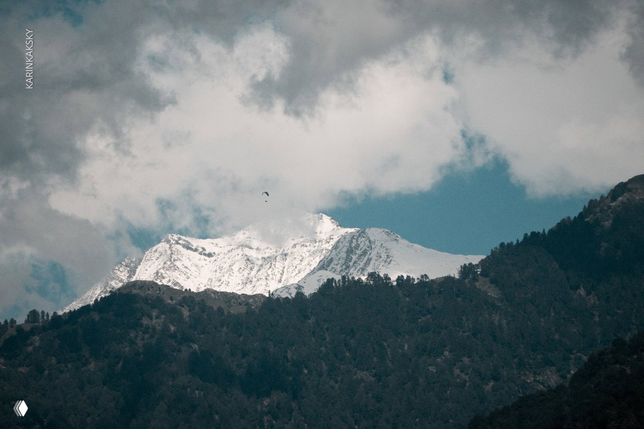 Вдалеке снежная вершина Гималаев и маленький параплан в небе над хребтом, подчёркивающий масштаб гор и полёта.