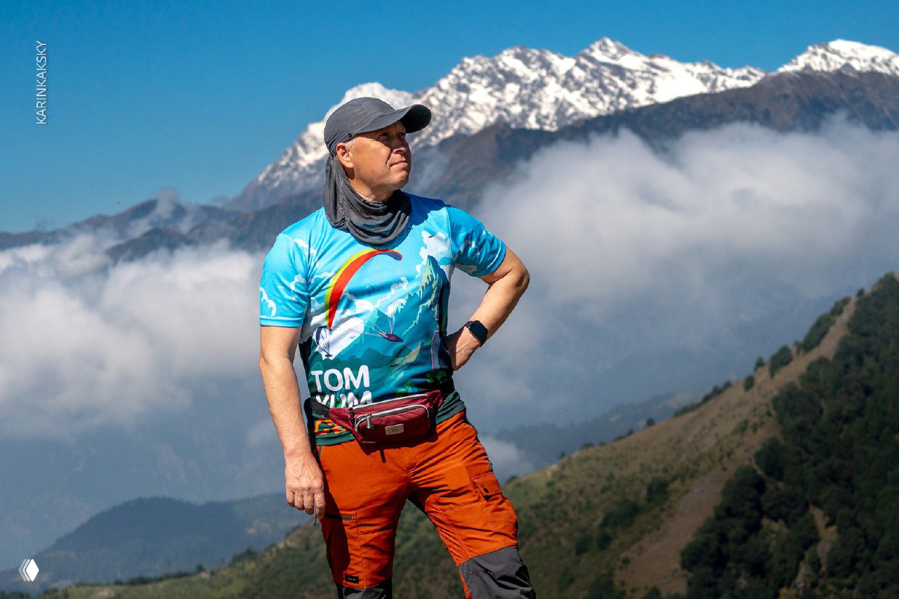 Пилот на фоне горного пейзажа в яркой футболке и снаряжении, готовый к полёту у снежных гималайских вершин.
