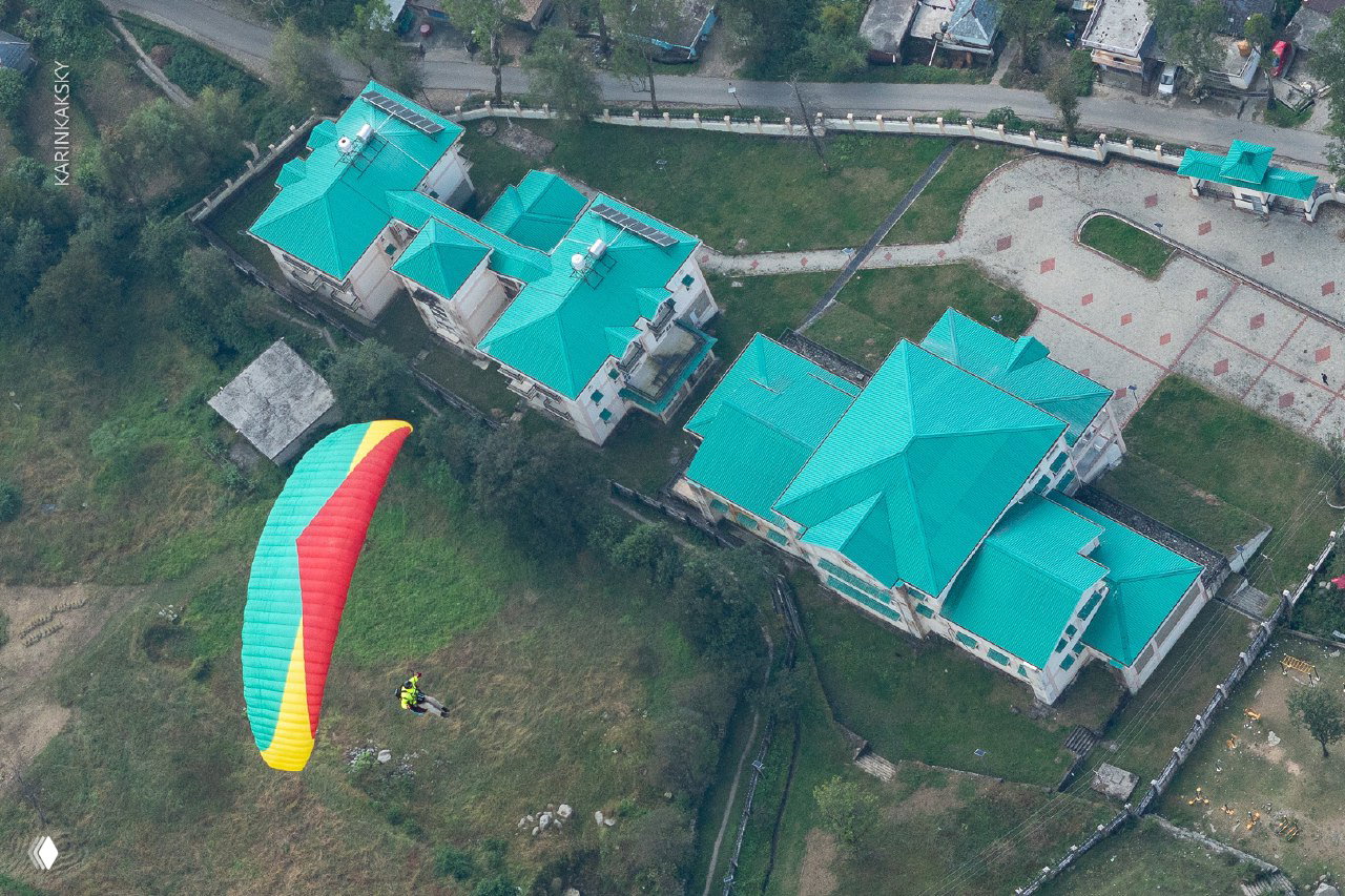 Вид сверху: яркий параплан летит над зелёной застройкой с бирюзовыми крышами и посадочной зоной внизу.
