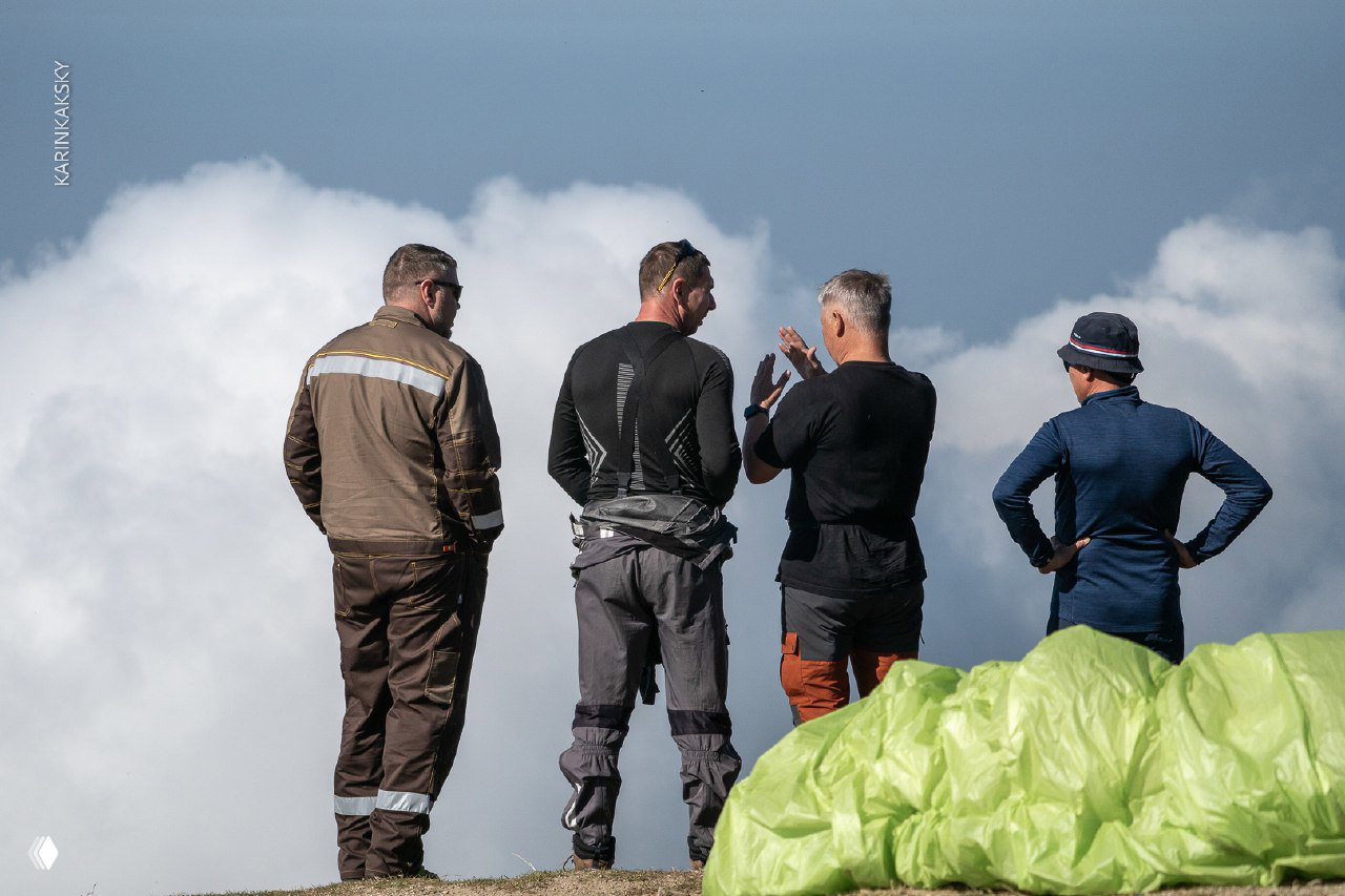 Группа пилотов обсуждает погоду и маршрут на краю старта, облачный фон и подготовленный параплан в переднем плане.