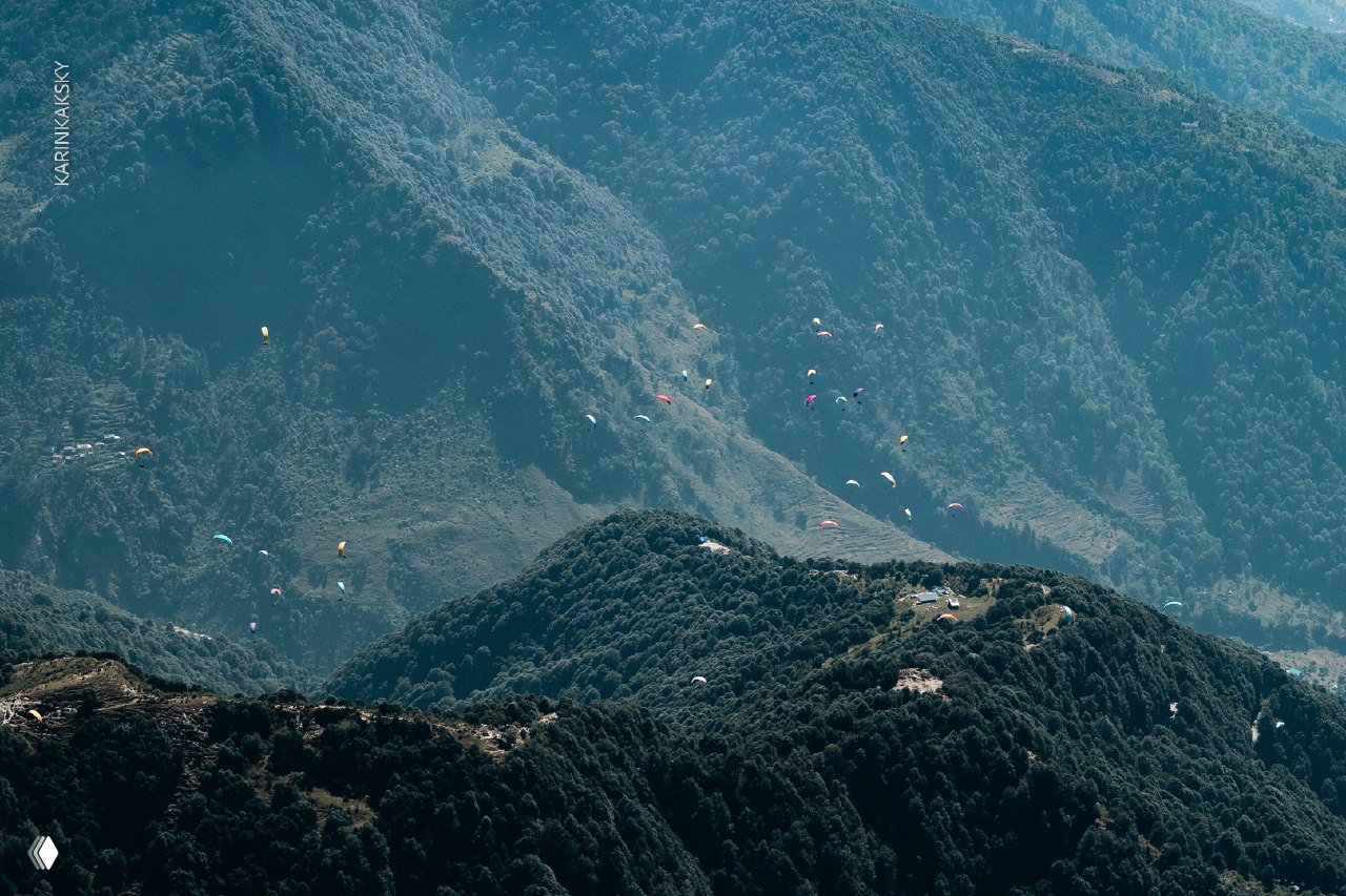 Общий вид хребта с множеством парапланов на фоне лесистых склонов и рассредоточенных пилотов по маршруту в долине.