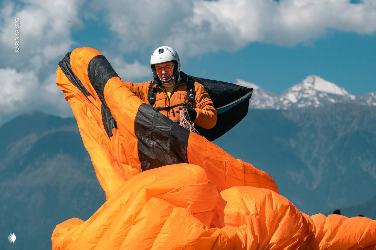 Пилот сворачивает или расправляет яркий оранжевый параплан на фоне гор и неба; крупный план подготовки экипажа перед полётом.