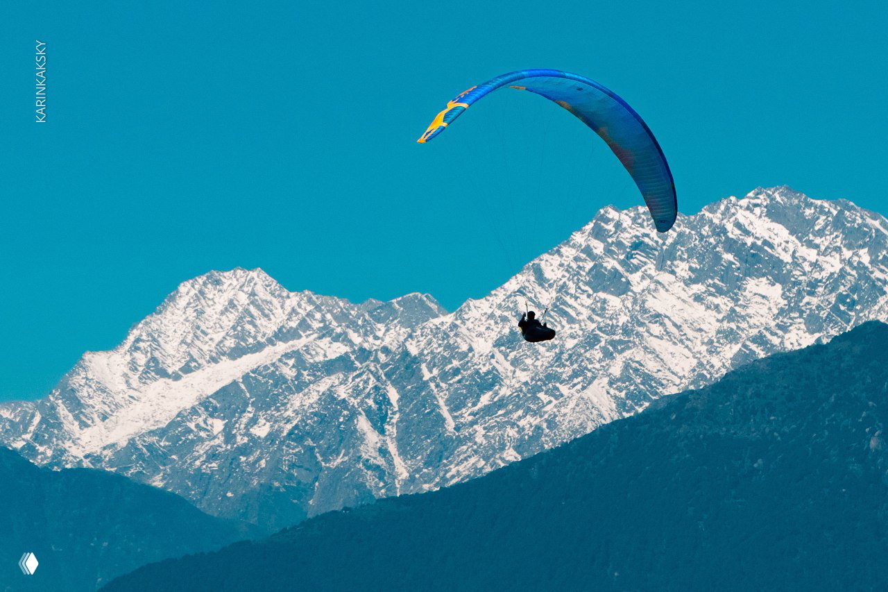 Одинокий пилот на синем куполе парит на фоне снежной горной стенки; контраст тёплого крыла и холодного горного рельефа.