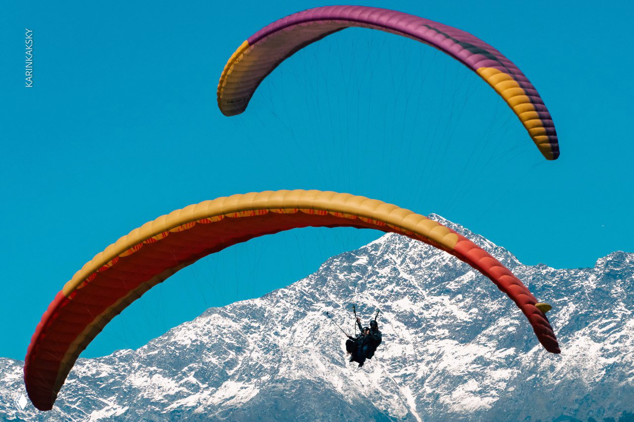 Два параплана на фоне заснеженных вершин: цветные купола в небе и силуэты пилотов на фоне голых горных склонов.