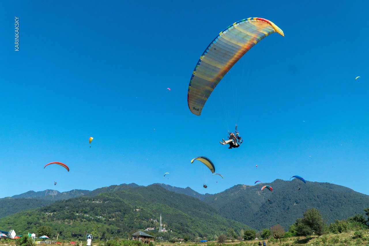 Группа парамоторных и парапланерных крыльев в ясном небесном пространстве над горным предгорьем и зелёными склонами.
