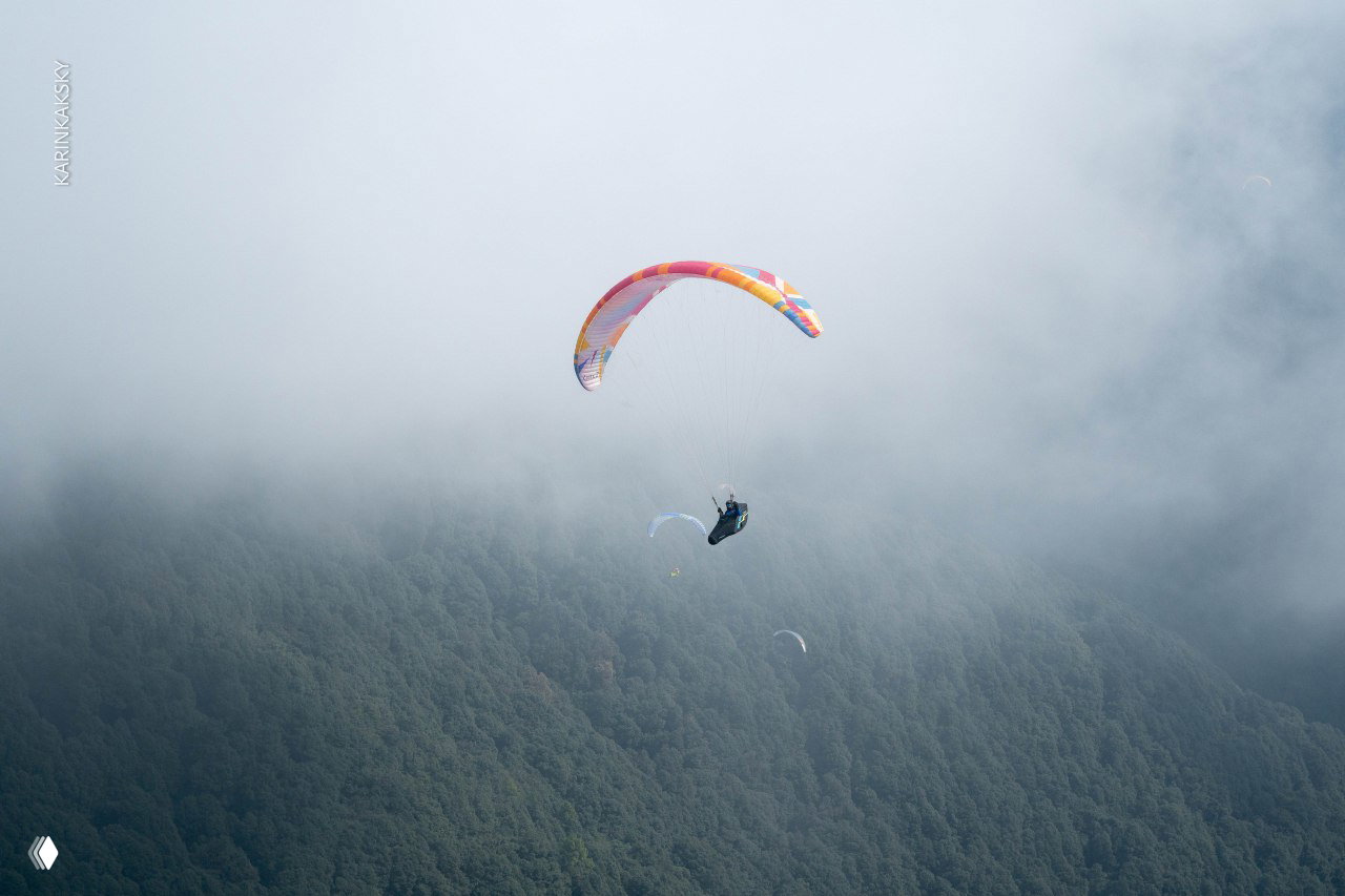 Парапланист в полёте над лесистым склоном, рядом сгущающиеся облачные пласты — иллюстрация того, как быстро меняется горная погода.