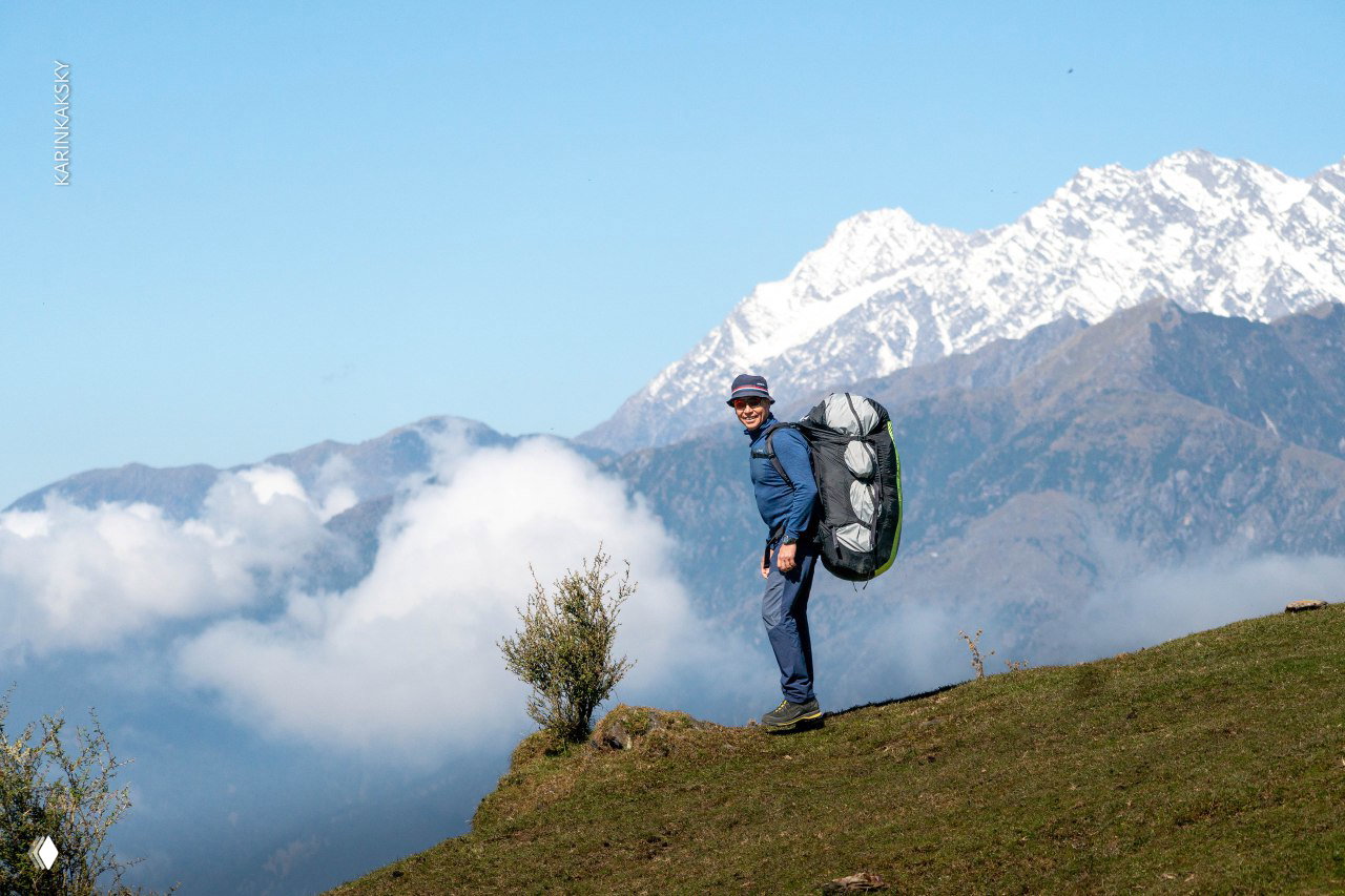 Улыбающийся пилот с рюкзаком-парапланом на фоне заснеженных вершин и облаков внизу — момент перед стартом в ясную, но изменчивую погоду.