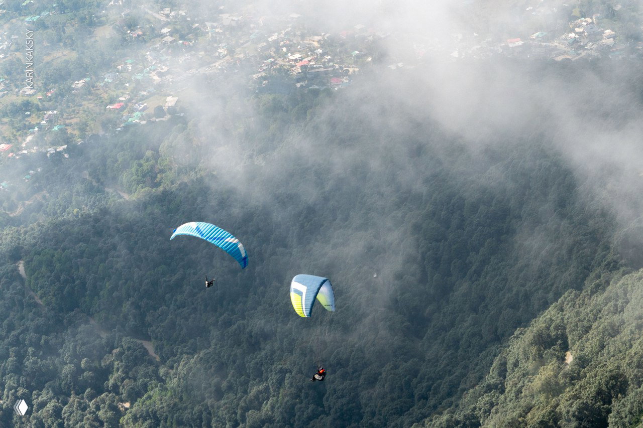 Два параплана маневрируют над густым лесом, между ними струятся облачные валы — сцена полётов в переменчивых горных условиях.