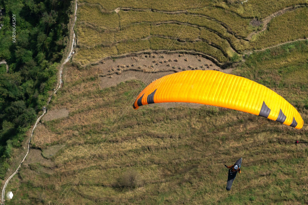 Крупный кадр ярко-оранжевого купола параплана над террасированными полями, вид сверху подчёркивает контраст между землёй и небом.