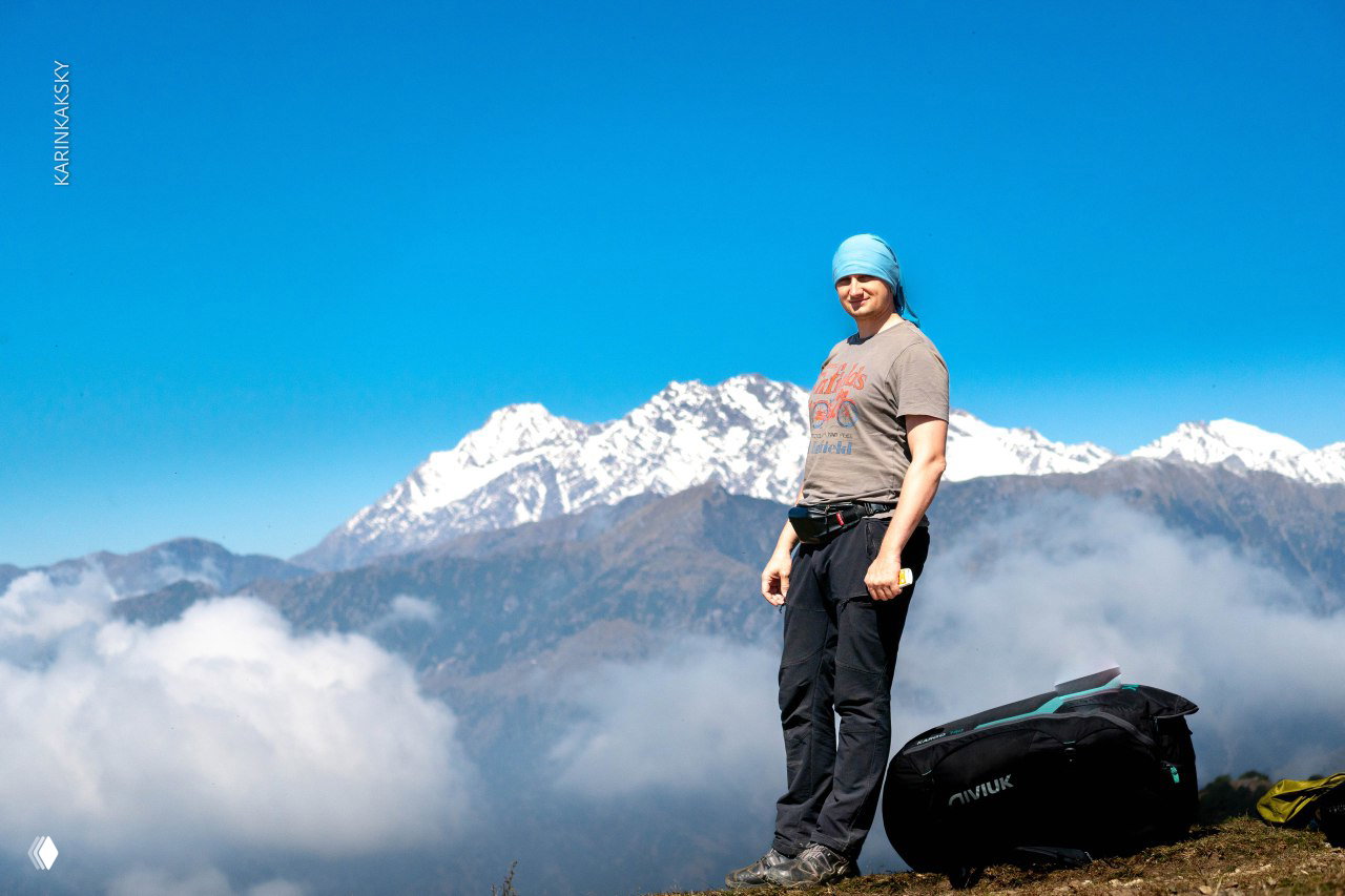 Пилот стоит на краю склона в ясную погоду, внизу облачные валы — момент ожидания и наблюдения за быстро меняющейся обстановкой.