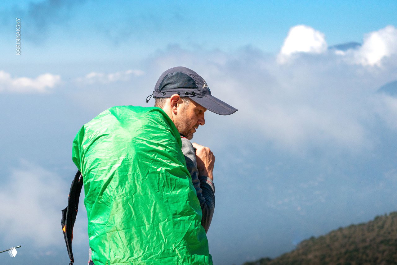 Крупный план пилота в зелёной защитной накидке, вокруг низкие облака и горный рельеф — деталь экипировки и атмосферная сцена перед полётом.