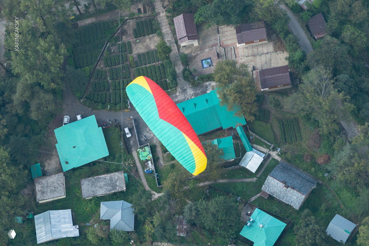 Вид сверху: яркий купол параплана в полёте над зелёной деревенской застройкой — динамика полёта и контраст природы и поселения.