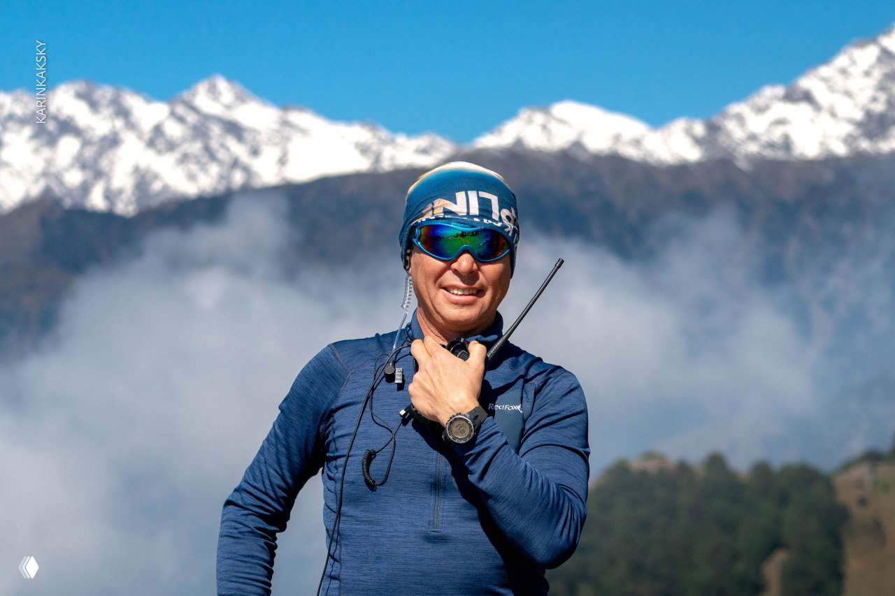 Пилот в очках и шапке улыбается на фоне гор и облаков; кадр передаёт настроение и связку людей с погодой и горами.