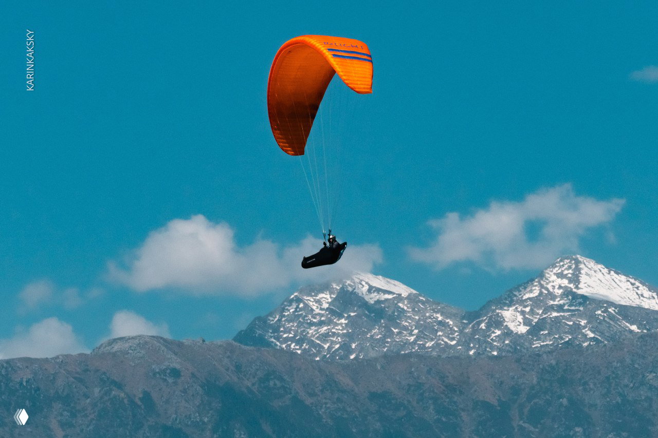 Оранжевый параплан висит над горными хребтами со снежными вершинами на фоне чистого голубого неба и небольших кучевых облаков.