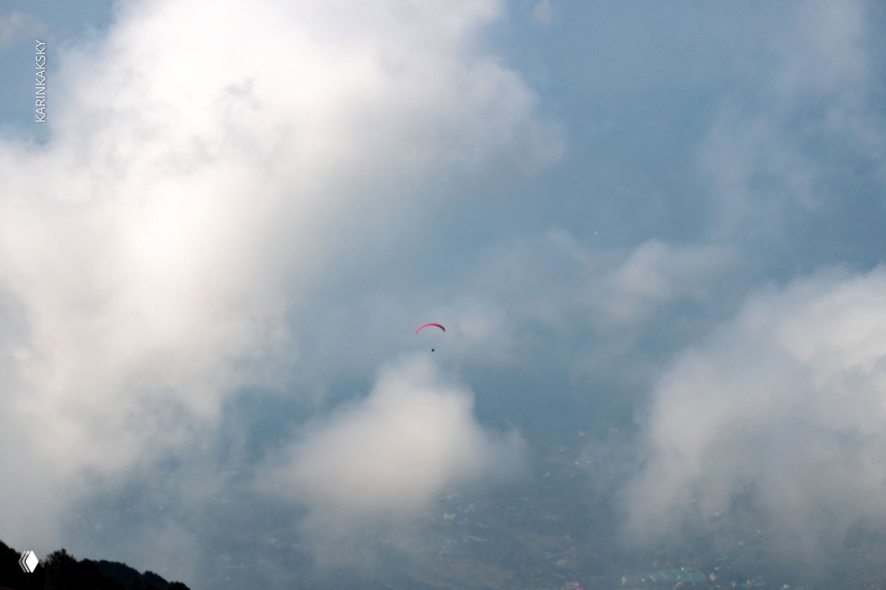 Маленький яркий параплан вдалеке среди крупных пышных облаков над горной долиной, создающий ощущение масштаба и высоты.